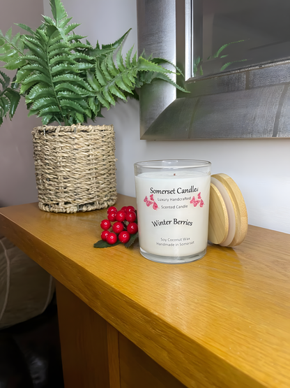 Winter Berries Scented soy candle displayed on a wooden mantel Piece with berries in the back ground. The candle is white in colour with bright red butterflies fluttering above the scent name. A bamboo lid is lent against the right side of the candle