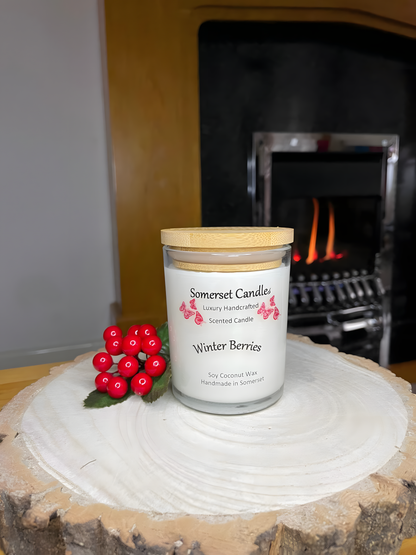 Winter Berries Scented soy candle displayed on a wooden board with berries. There is a lit fire in the back ground. The candle is white in colour with bright red butterflies fluttering above the scent name and fitted with a lovely bamboo lid.