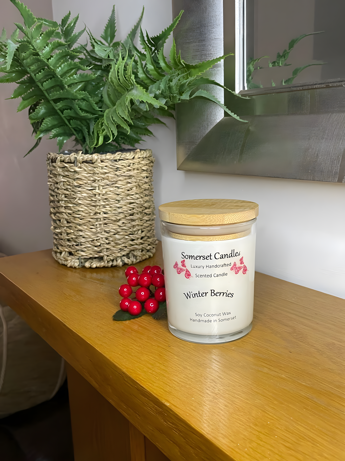 Winter Berries Scented soy candledisplayed on a wooden mantel piece with berries in the back ground. The candle is white in colour with bright red butterflies fluttering above the scent name. A bamboo lid is fitted to the candle.