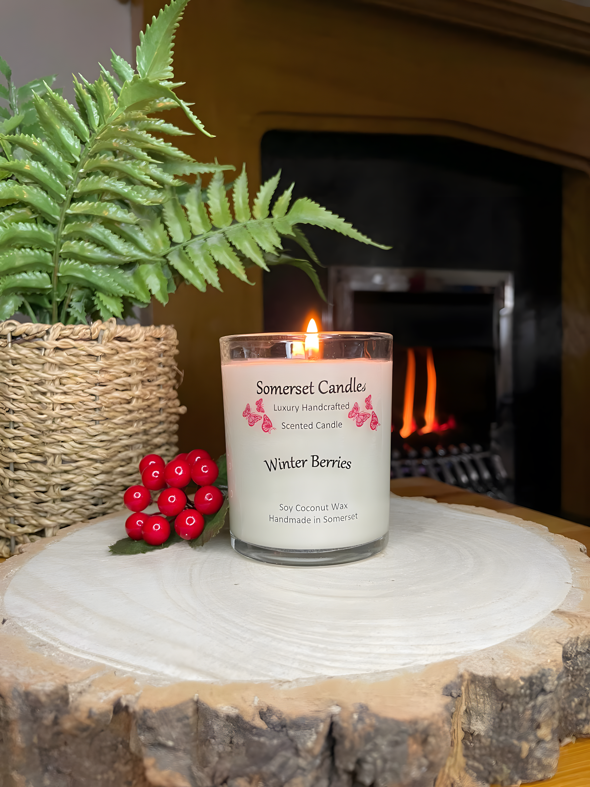 Winter Berries Scented soy candle displayed on a wooden board with berries. The candle is lit, is white in colour and has bright red butterflies fluttering above the scent name. There is a fire in the back ground.