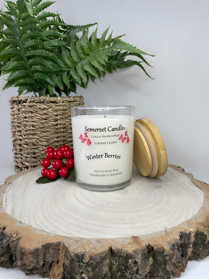 Winter Berries Scented soy candle displayed on a wooden board with berries in the back ground. The candle is white in colour with bright red butterflies fluttering above the scent name. A bamboo lid is lent against the right side of the candle.