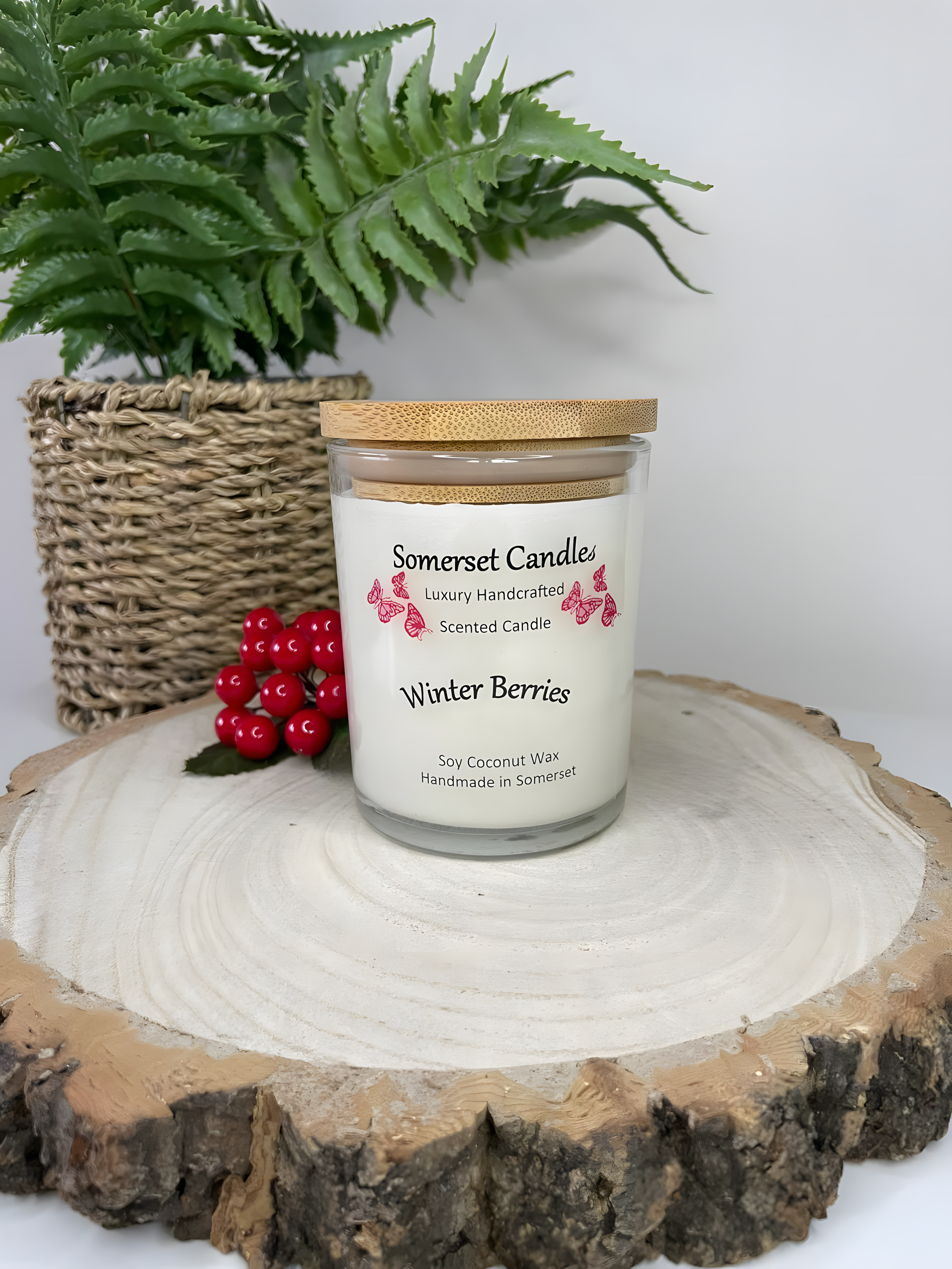 Winter Berries Scented soy candle displayed on a wooden board with berries in the back ground. The candle is white in colour with bright red butterflies fluttering above the scent name and fitted with a lovely bamboo lid.