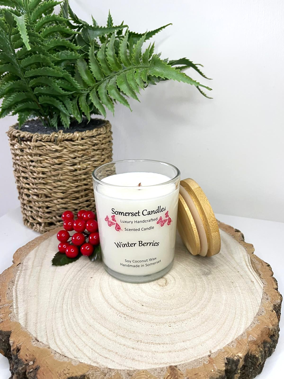 White soy candle in a 7oz clear glass jar with a natural wooden lid. The candle is made from a soy-coconut blend and scented with a fruity scent of winter berries. Above the scent name on the label, several red butterflies are displayed, adding a romantic touch. Clean, minimalist design ideal for gifts, home decor or self-care.