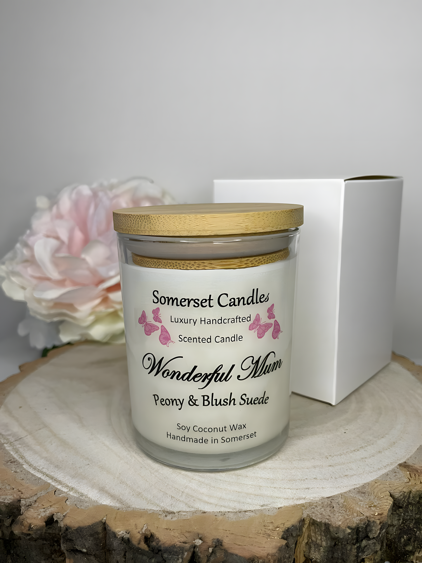 Wonderful Mum Peony & Blush Suede Soy Candle displayed on a wooden board. Behind is a white gift box and a beautiful pink peony. The candle is white in colour with bright pink butterflies fluttering above the scent name and fitted with a lid.