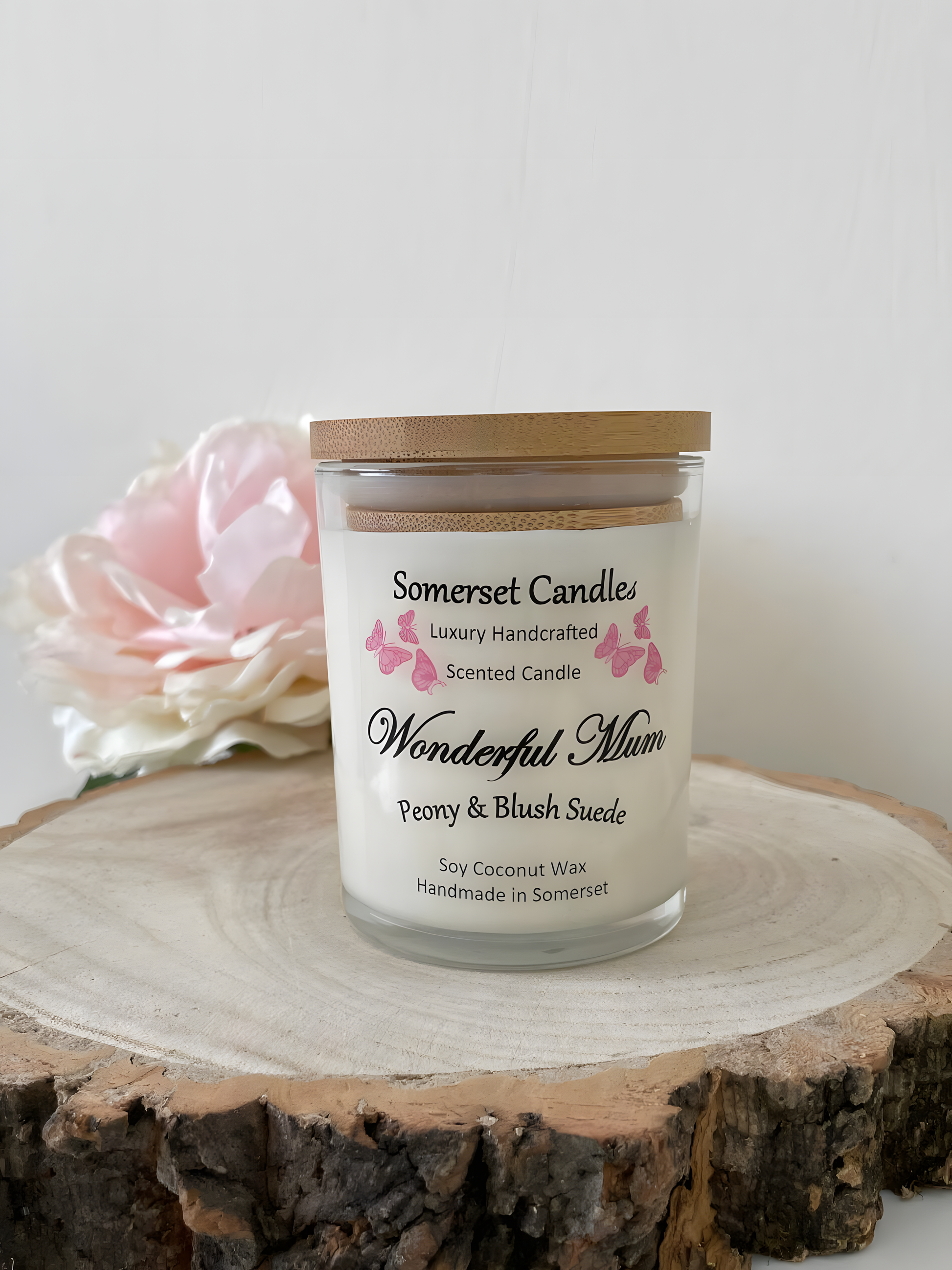 Wonderful Mum Peony & Blush Suede Soy Candle wooden board with a beautiful pink peony behind. The candle is white in colour with bright pink butterflies fluttering above the scent name and fitted with a lovely bamboo lid.