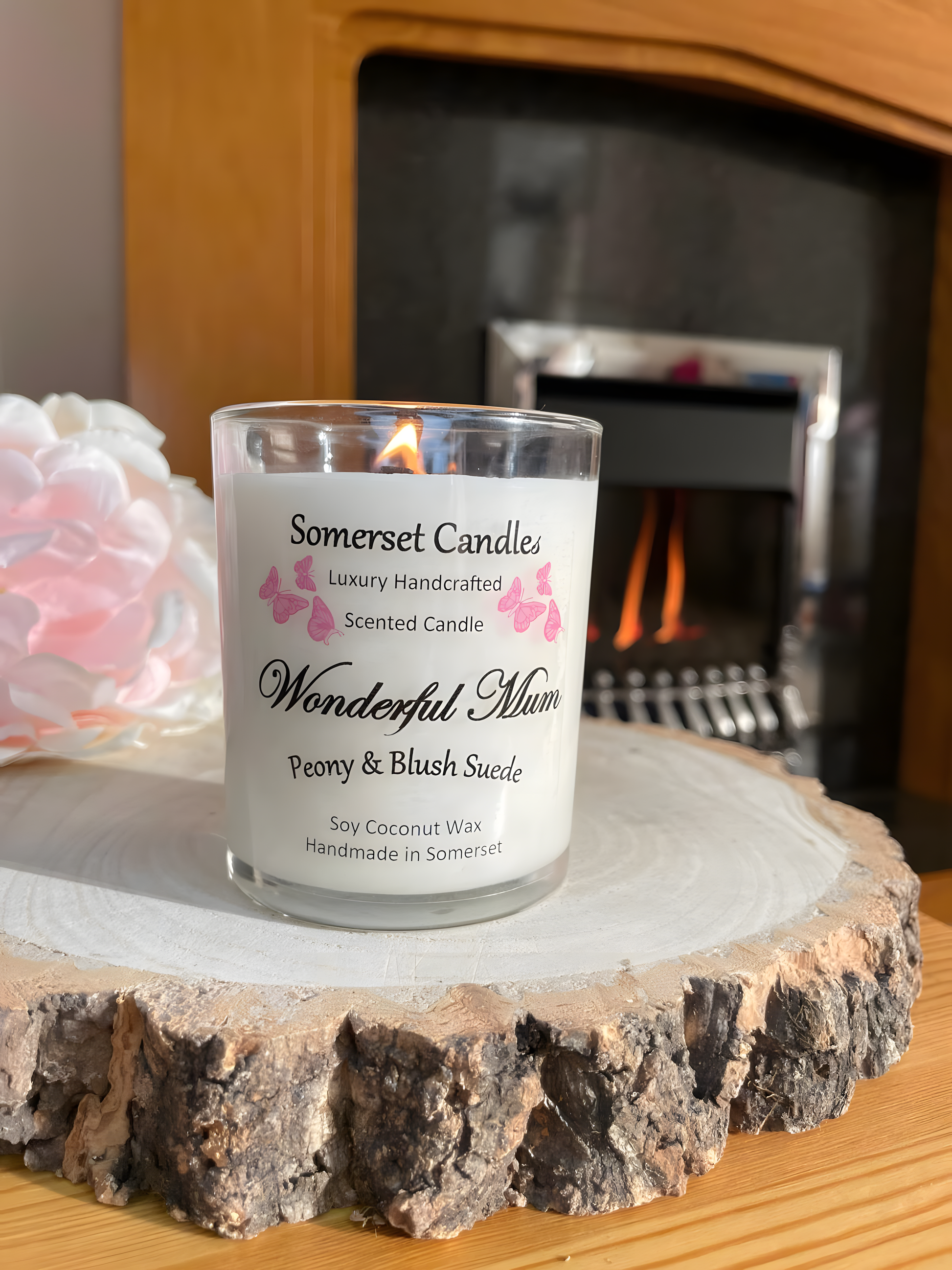 Wonderful Mum Peony & Blush Suede Soy Candle is displayed on a wooden board with a beautiful pink peony behind. The candle is white in colour with bright pink butterflies fluttering above the scent name. The candle is lit and in front of a fire.