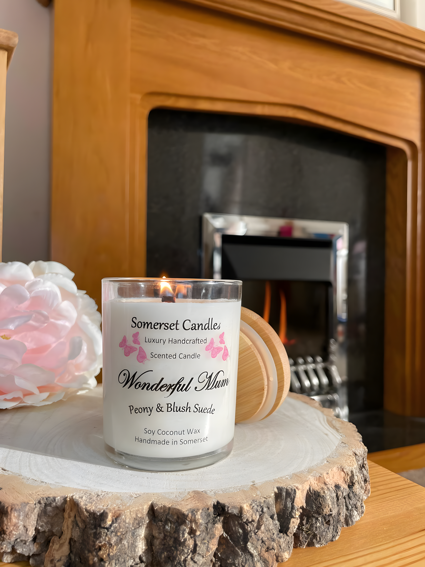 Wonderful Mum Peony & Blush Suede Soy Candle is displayed on a wooden board with a beautiful pink peony behind. The candle is white in colour with bright pink butterflies fluttering above the scent name. The candle is lit and in front of a fire.