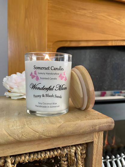 Wonderful Mum Peony & Blush Suede Soy Candle displayed in front of a fire with a beautiful pink peony behind. The candle is white in colour with bright pink butterflies fluttering above the scent name and with a bamboo lid to one side.