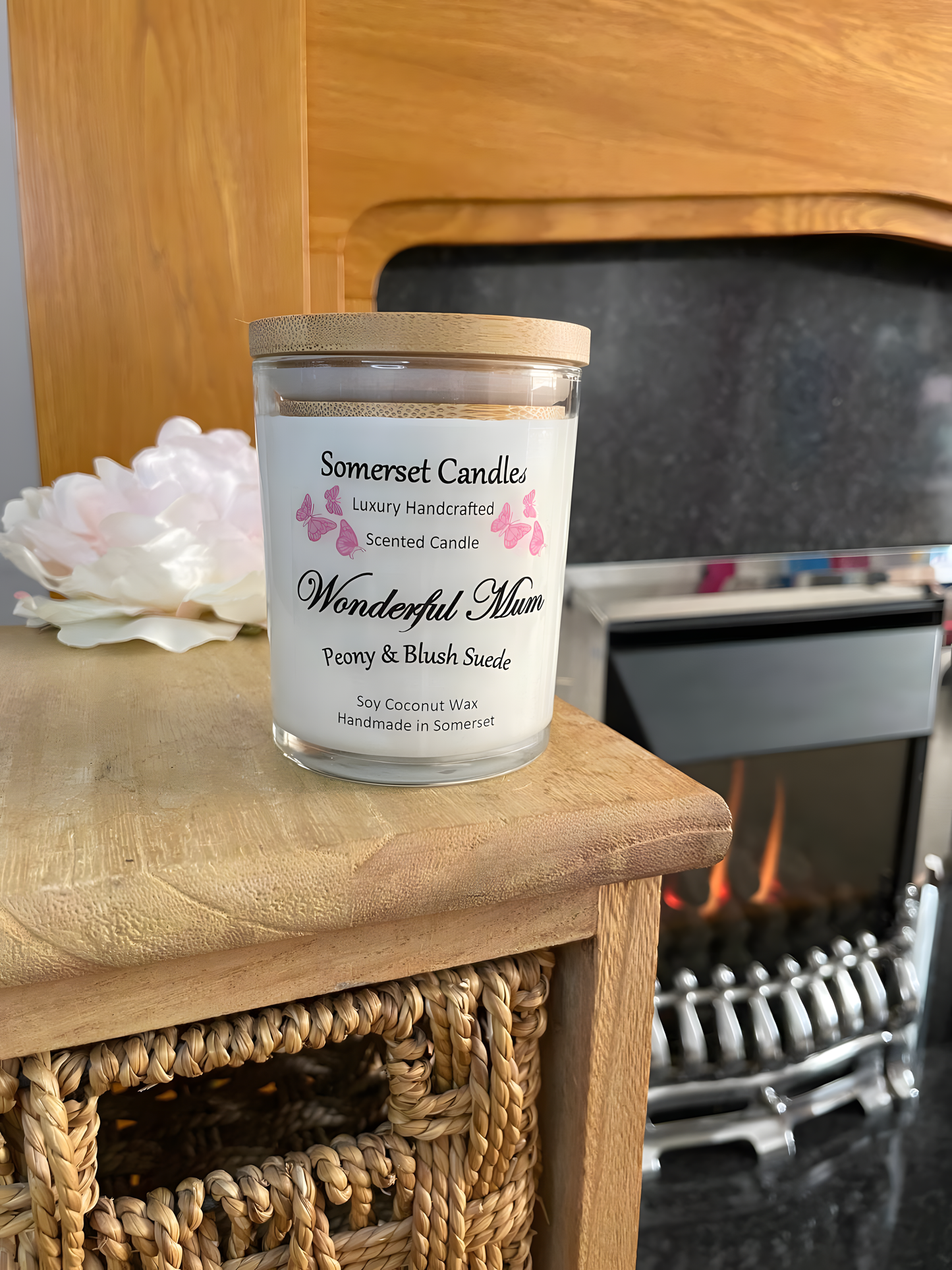 Wonderful Mum Peony & Blush Suede scented soy candle displayed in front of a fire with a beautiful pink peony behind. The candle is white in colour with bright pink butterflies fluttering above the scent name and fitted with a lovely bamboo lid.