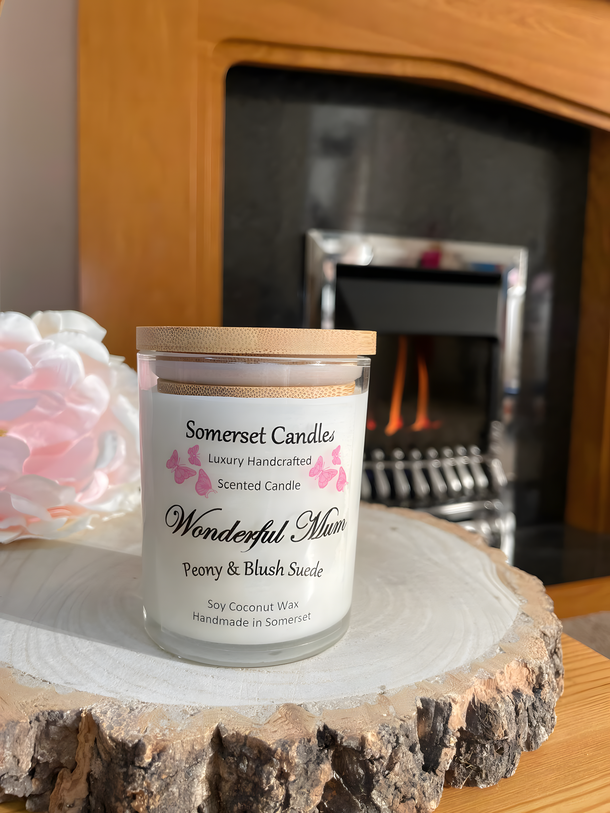 Wonderful Mum Peony & Blush Suede Soy Candle  displayed in front of a fire with a beautiful pink peony behind. The candle is white in colour with bright pink butterflies fluttering above the scent name and fitted with a lovely bamboo lid.