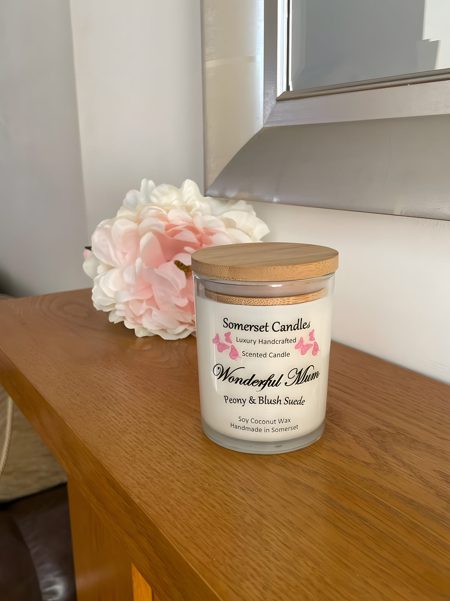 Wonderful Mum Peony & Blush Suede Soy Candle is displayed on a wooden mantel piece with a beautiful pink peony behind. The candle is white in colour with bright pink butterflies fluttering above the scent name and fitted with a lovely bamboo lid