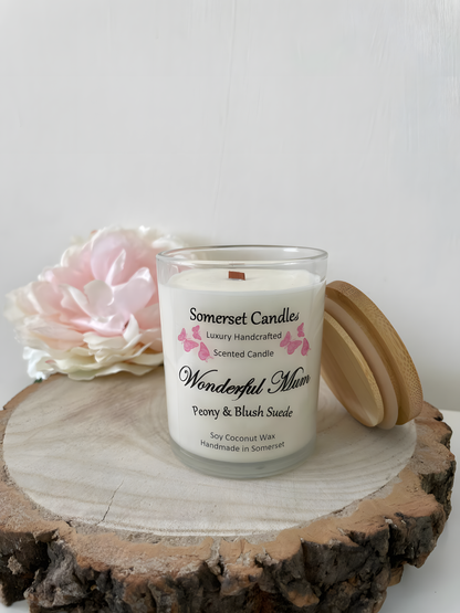 Wonderful Mum Peony & Blush Suede is displayed on a wooden board with a beautiful pink peony behind. The candle is white in colour with bright pink butterflies fluttering above the scent name. The lid is resting on the right side.