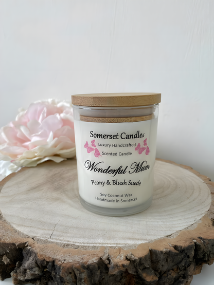Wonderful Mum Peony & Blush Suede Soy Candle is displayed on a wooden board with a beautiful pink peony behind. The candle is white in colour with bright pink butterflies fluttering above the scent name and fitted with a lovely bamboo lid.